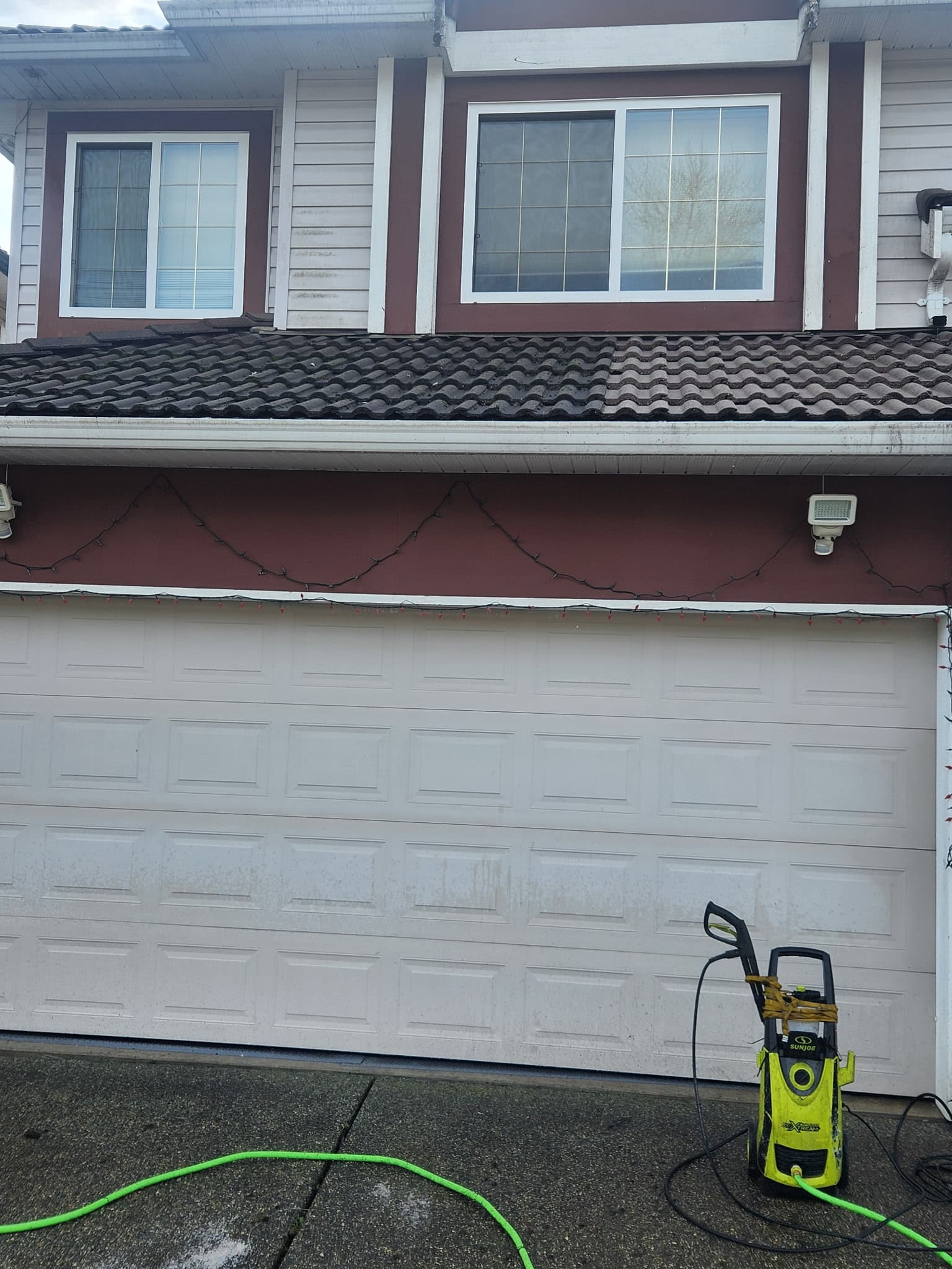 Pressure washer on a driveway in front of a house with partially cleaned roof tiles.